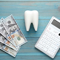 A ceramic tooth model, hundred-dollar bills, and a calculator on a light blue wooden table
