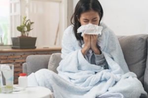 Woman sitting on her couch with a blanket over her shoulders as she sneezes into a tissue