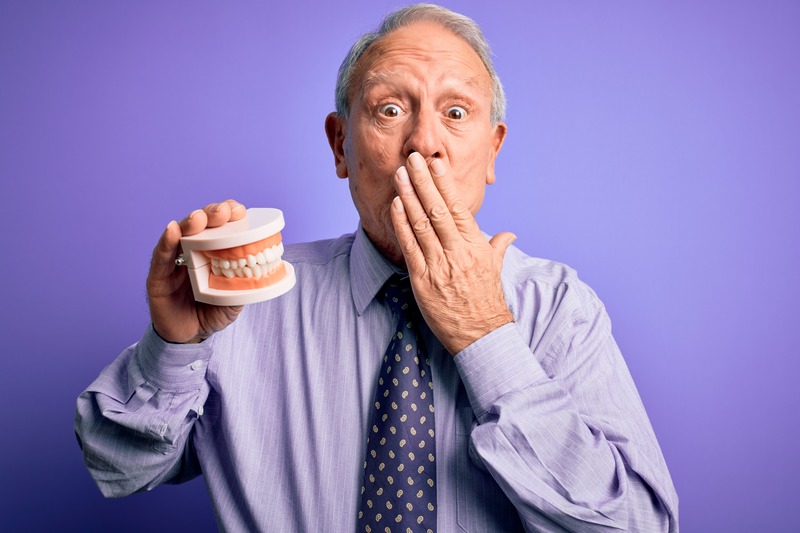 A gray haired man holding prosthetic dentures while covering his mouth